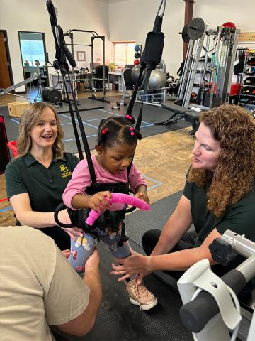 Two women work with a young girl that is strapped into a suspended harness during a physical therapy session. 
