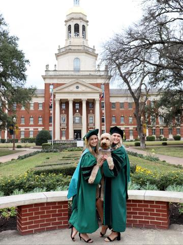 Two women, in graduation gowns, pose with their dog in front of Baylor University's Pat Neff Hall. 